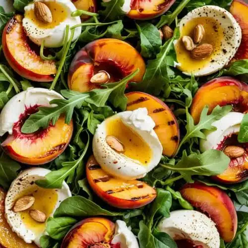 A high-quality food photograph of a fresh summer salad on a white ceramic plate. The salad consists of grilled peach slices with charred grill marks, sliced white burrata cheese with oozing cream, and fresh arugula leaves. The peaches are a vibrant orange-red color with deep red blushes, cut into thick circular slices. The burrata cheese is perfectly circular, showing the distinctive white outer layer and creamy interior. Small whole almonds are scattered throughout the salad, with some resting on top of the cheese. The arugula leaves are bright green and provide a fresh, peppery contrast to the sweet peaches. The salad is photographed from directly above on a white textured surface. The lighting is bright and natural, highlighting the glossy surface of the grilled peaches and the creamy texture of the cheese. The composition is filled with the ingredients, creating a dense, colorful arrangement that fills the entire frame.