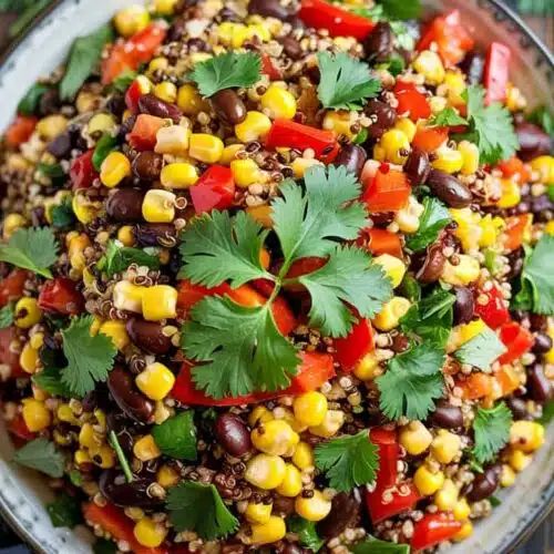 A vibrant Mexican-style quinoa salad photographed from above on a dark gray ceramic plate. The salad contains black beans, yellow corn kernels, diced red bell peppers, and fresh green cilantro leaves scattered throughout. Large fresh cilantro sprigs are placed decoratively on top of the salad. Two lime wedges are positioned at the top right corner of the frame, with their bright green flesh visible. The salad is served on a rustic dark wooden surface. A blue-gray linen napkin is partially visible in the bottom left corner. The lighting is bright and natural, highlighting the various textures and colors of the ingredients. The composition is shot in a square format with professional food photography styling.